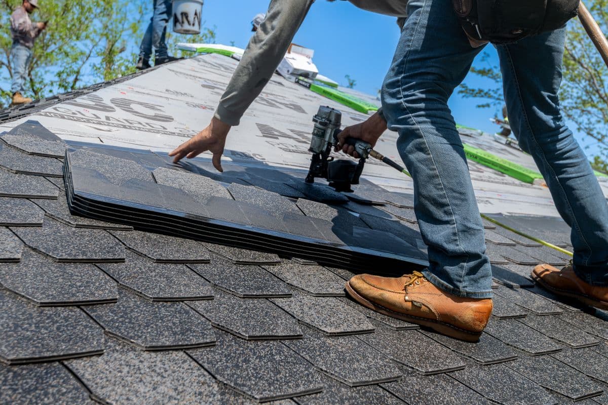 A roofer kneeling on a rooftop using a nail gun to fasten new asphalt shingles