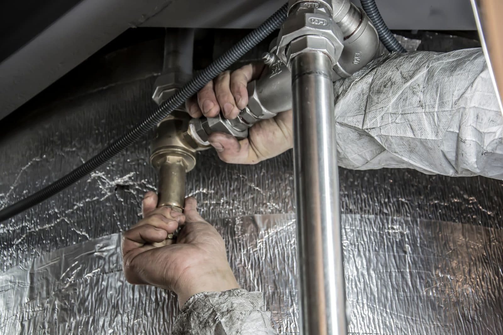 Plumber's gloved hands fitting brass and steel supply pipes under insulation