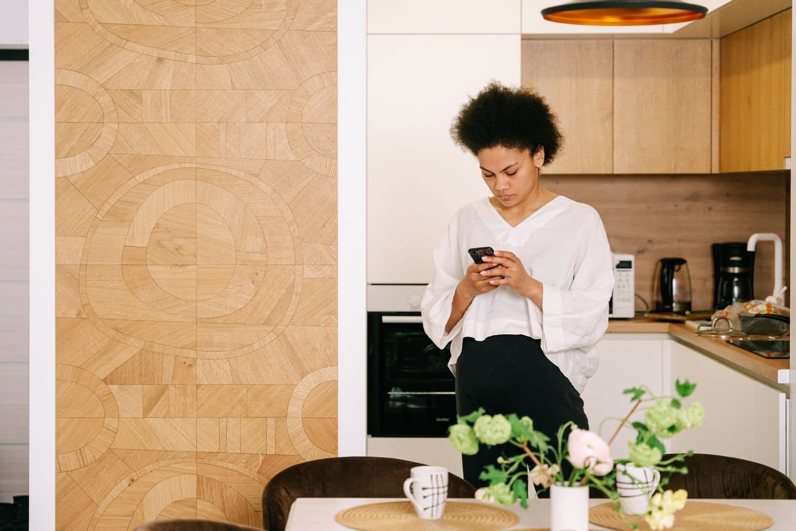 A woman standing in her kitchen looking down at her phone, a dining table with flowers in the foreground