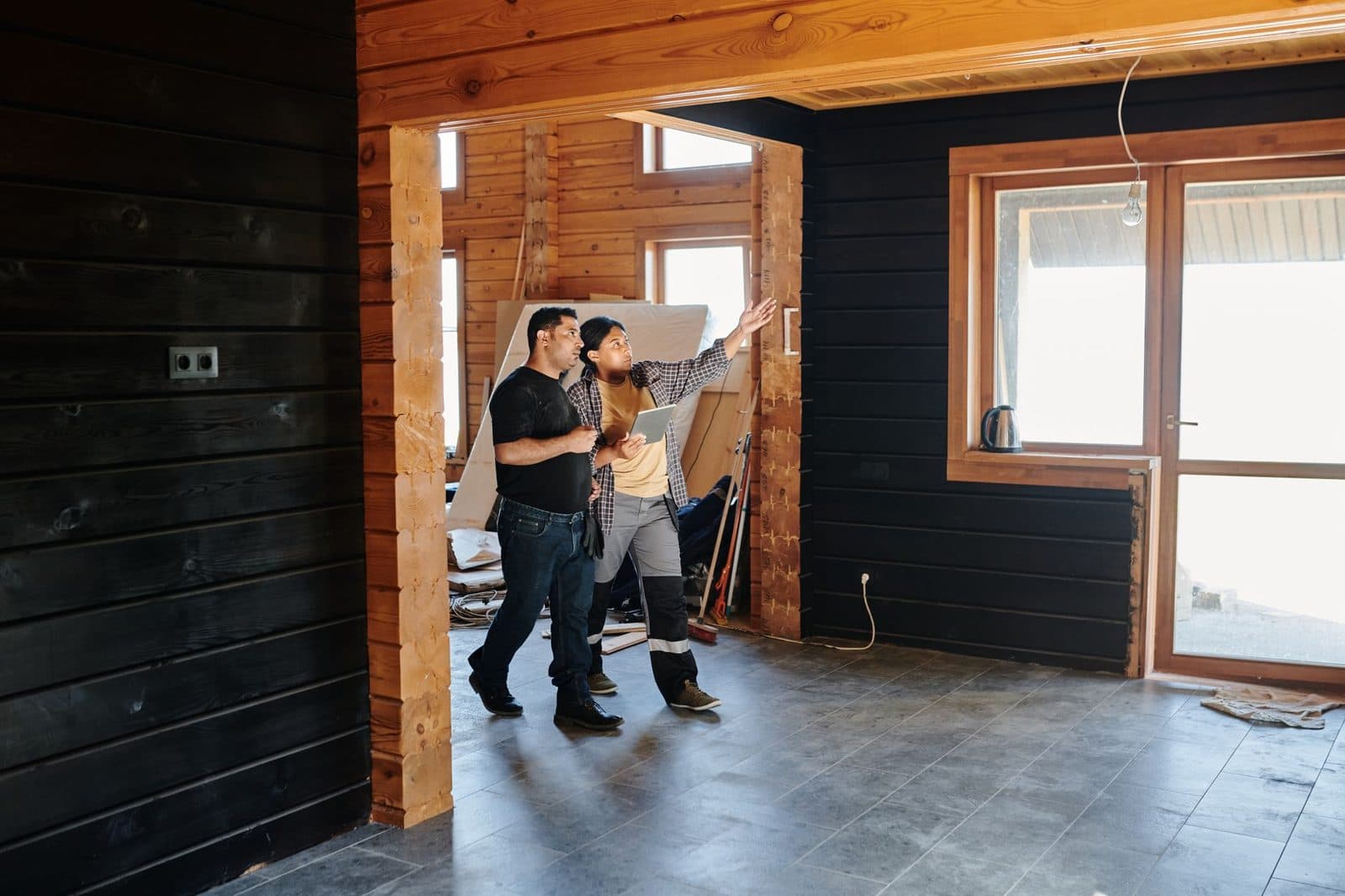 A contractor holding a tablet walks a homeowner through a partly finished room, gesturing up at the wall