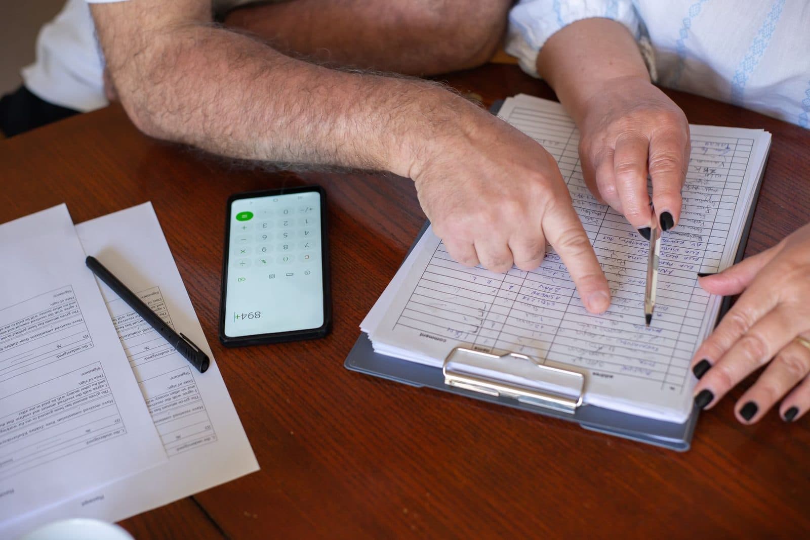 Two people at a wooden table going through an itemized handwritten estimate on a clipboard, with a calculator and a signature form beside it