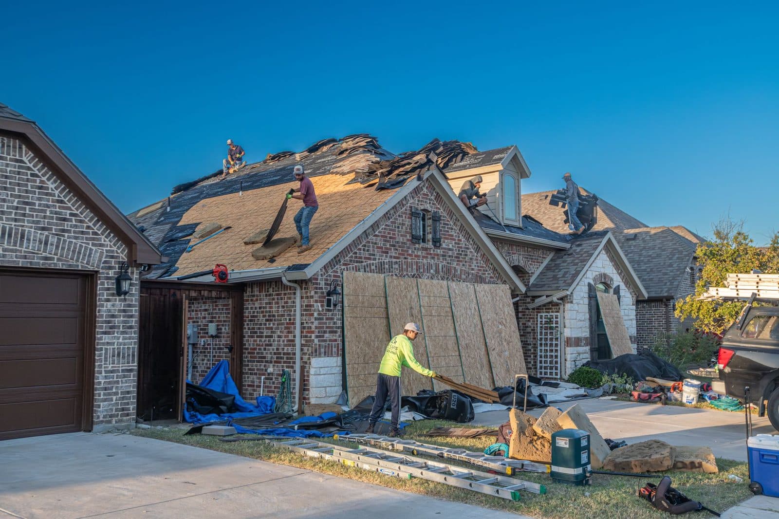 A five-person roofing crew mid tear-off on a two-story brick house, old shingles stacked on the roof and materials staged in the driveway